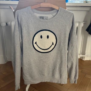 Grå sweatshirt med smiley från Dedicated - Ljusgrå sweatshirt från Dedicated med ett stort broderat smiley-ansikte på bröstet. Är 173cm lång och den sitter bra vid midjan, inte så lång! Kan mötas i sthlm (klicka INTE köp nu, skriv privat ist!)