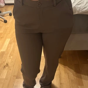 Bruna kostymbyxor  - Snygga bruna kostymbyxor med utsvängda / raka ben och medel-hög midja. Byxorna har bälteshällor, knapp och dragkedja framtill. Perfekta till en stilren look och passar till många olika tillfällen. Oanvänd!