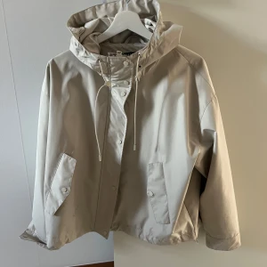 Beige vindjacka med huva från ZARA - Beige vindjacka från ZARA med huva. Jackan har knappar framtill, två fickor med knapp samt justerbara ärmslut. 