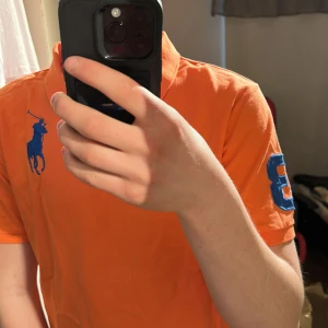 Orange pikétröja från Ralph Lauren - Säljer en orange pikétröja från Ralph Lauren med blå logga på bröstet och en blå siffra 3 på ärmen. Klassisk krage och korta ärmar. Perfekt för en sportig och stilren look. Nya taggen 
