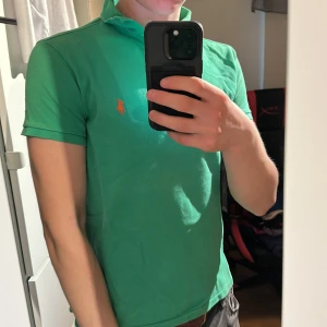 Grön pikétröja från Ralph Lauren - Säljer en klassisk grön pikétröja från Ralph Lauren med orange broderad logga på bröstet. Tröjan har krage, knappar vid halsen och korta ärmar. Perfekt för en stilren och sportig look. Gamla taggen. M men lite mer som s 