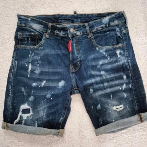Mörkblå jeansshorts Dsquared2 M/46 - Snygga mörkblåa Dsquared2 jeansshorts med slitna detaljer, färgstänk och upprullade benslut. Klassisk femficksmodell med knappgylf och en stor patch med Dsquared2-logga baktill. Perfekta för en avslappnad streetstil.