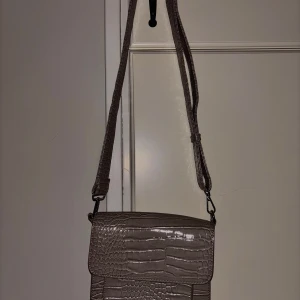 Beige axelväska i krokopräglad syntet från HVISK - Säljer en beige axelväska från HVISK med krokopräglad yta och bred axelrem. Väskan har magnetlås och stilren design, perfekt för att lyfta din outfit. Rymmer det viktigaste och är lätt att matcha.