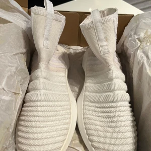 Vita Adidas sneakers med stickad ovandel - Säljer ett par vita Adidas sneakers med stickad ovandel och högt skaft. Skorna har en unik ribbad design och är tillverkade i syntetmaterial. Perfekta för dig som gillar en modern och sportig look.