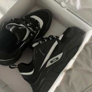 Svarta D1or sneakers med vita detaljer - Säljer ett par svarta D1or sneakers med vita och grå detaljer. Skorna har en chunky sula, meshpartier och snörning framtill. Märket syns tydligt på sidan. Perfekta för dig som gillar exklusiva och trendiga sneakers.