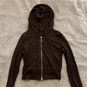 Brun croppad hoodie med dragkedja - Säljer en brun croppad hoodie med dragkedja framtill och huva. Jackan har smala vertikala ränder och långa ärmar. Perfekt för en avslappnad och trendig look.