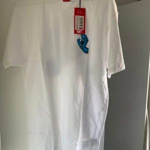 Vit t-shirt från Santa Cruz med blå handtryck - Säljer en vit t-shirt från Santa Cruz med ikoniskt blått handtryck på bröstet och ryggen. Klassisk passform och rund halsringning. Perfekt för dig som gillar streetwear och skatekultur.