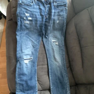 Blå slitna jeans Slim/Glenn - Säljer ett par blå jeans i modellen Slim/Glenn med slitna detaljer och snygga slitningar framtill. Jeansen har klassisk femficksdesign och kontrastsömmar på bakfickorna. Passformen är slim och storleken är 31/32.