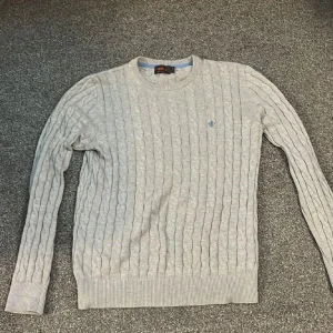 Ljusgrå kabelstickad tröja från Morris - Säljer en ljusgrå kabelstickad tröja från Morris i 100% pima cotton. Tröjan har rund halsringning, lång ärm och en liten blå logga broderad på bröstet. Perfekt för lager-på-lager och stilren look. Pris kan diskuteras vid snabb affär.