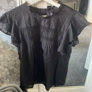 Svart blus med volang från Vero Moda - Säljer en svart blus från Vero Moda med korta volangärmar. Blusen har rynkade partier framtill och en knapp i nacken. Perfekt för dig som gillar en luftig och feminin stil. De står att de är storlek L men jag har S och den sitter bra på mig❤️