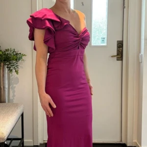 Sericerosa långklänning med twistad överdel - Elegant sericerosa långklänning med twistad detalj och liten öppning vid bysten. Klänningen har korta ärmar och figurnära passform som framhäver siluetten. Perfekt för speciella tillfällen.