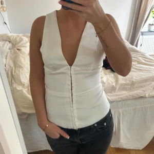 Vit djup v-ringad topp - Säljer en vit ärmlös topp med djup v-ringning och figurnära passform. Toppen har knäppning framtill och är perfekt för dig som vill ha en stilren och trendig look.