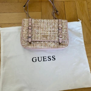 Tweed axelväska från Guess - Säljer en snygg axelväska från Guess i tweed med pastellfärger och ljusrosa detaljer. Väskan har guldfärgade kedjor, dubbla spännen framtill och logotyp i metall. Dustbag tillkommer. Banden går att justera från crossbody till axelrem. Använd 1 gång