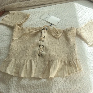 Beige offshoulder blus med volang och knytning från Zara - Superfin beige offshoulder blus från Zara med volangkant nertill och snörning framtill. Blusen har korta ärmar och är gjord i ett lätt, strukturerat material som passar perfekt till sommaren.