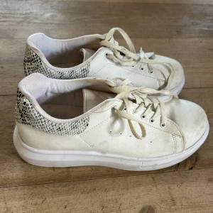 Vita sneakers med ormmönstrad detalj - Säljer ett par vita sneakers från My Shoes med snygg ormmönstrad detalj på hälen. Skorna har snörning och en tjock vit sula som ger en sportig look. Perfekta för dig som vill ha en stilren men ändå trendig sko.