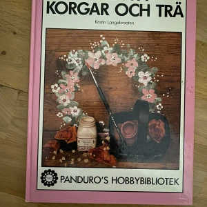 Bok om Måla på korgar och trä - En bok med inspiration och tips om hur du kan måla och dekorera spånkorgar och träföremål. Omslaget är rosa med bilder på målade blommor, penslar och färg. Perfekt för dig som gillar pyssel och DIY-projekt. Nyskick 
