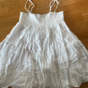 Vit somrig klänning med smock och tunna axelband - Säljer en luftig vit klänning med smock upptill och tunna axelband. Klänningen har volanger, säljer då den är lite för liten för mig💞