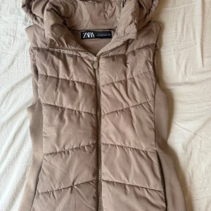 Beige quiltad dunväst från Zara - Säljer en beige quiltad dunväst från Zara med huva och dragkedja framtill. Västen har en sportig look och är perfekt för lager-på-lager-stil under kyligare dagar.