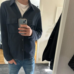 Mörkblå overshirt från Zara - Säljer en stilren mörkblå overshirt från Zara med klassisk krage och fyra stora fickor framtill. Jackan har knäppning med knappar och är långärmad. Perfekt för lager-på-lager och enkel att matcha till jeans.