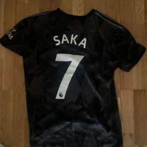 Arsenal bortatröja Saka 7 - Snygg svart Arsenal bortatröja från 2022/23 säsongen med Saka på ryggen. Tröjan är i fin fint skick och passar S och möjligtvis M!