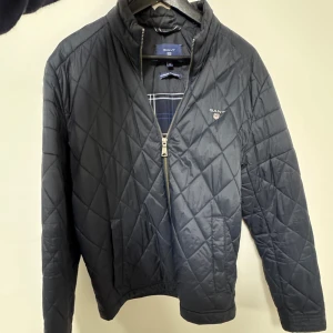Gant Svart quiltad jacka  - Snygg svart quiltad jacka från Gant med dragkedja framtill och diskret logga på bröstet. Jackan har ståkrage och ett stilrent rutmönster. Perfekt för dig som vill ha en klassisk och tidlös look.