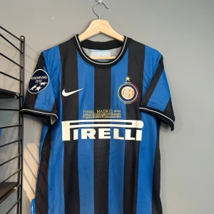 Inter Milan fotbollströja Sneijder 10 - Inter Milan matchtröja med Sneijder 10 på ryggen. Svart och blå vertikala ränder, Champions 2010-märke på ärmen och Pirelli som sponsor på bröstet. Broderad klubbmärke och Nike-logga framtill. Helt ny med tagg en replika i S