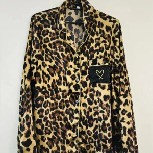 Leopardmönstrad pyjamasskjorta i satin - Säljer en leopardmönstrad långärmad skjorta i glansigt satinmaterial. Skjortan har svarta detaljer, knappar framtill och en svart bröstficka med ett broderat hjärta i gult. Perfekt för dig som vill sticka ut med ett djurmönster.