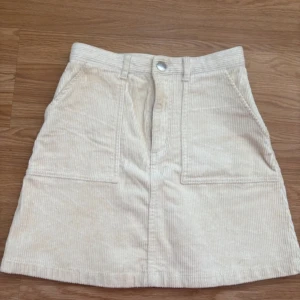 Beige manchesterkjol från Monki - Säljer en beige manchesterkjol från Monki med två stora fickor framtill och knappstängning i midjan. Kjolen har bälteshällor och är i en klassisk A-linjeformad modell. 