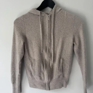 Beige hoodie i kashmir från Soft Goat - Mysig beige hoodie i kashmir från Soft Goat. Tröjan har dragkedja framtill, huva med dragsko och två fickor. Perfekt för en avslappnad och stilren look.