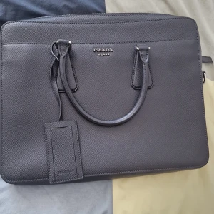 Svart portfölj från Prada - Säljer en stilren svart portfölj från Prada Milano. Väskan är tillverkad i exklusivt skinn med två handtag och en elegant, strukturerad yta. Perfekt för att bära laptop eller dokument. Klassisk design med diskret logga framtill.