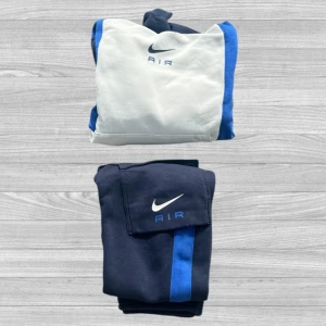 Nike Air tracksuit - Nike Air set med hoodie och byxor. Hoodien är vit med blå och svarta paneler, har huva och magficka. Byxorna är mörkblå med blå ränder och fickor med lock. Båda plaggen har Nike Air-logga broderad framtill.