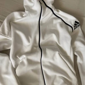 Vit hoodie från Adidas med svart dragkedja - Säljer en stilren vit hoodie från Adidas med svart dragkedja och Adidas-logga på ärmen. Jackan har huva och är långärmad, perfekt för träning eller chill. Klassisk sportig look med snygga kontrastdetaljer.