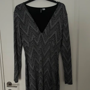 Svart och vit långärmad klänning med mönster från H&M - Säljer en svart långärmad klänning från H&M med vit, grafisk zigzag-mönster och djup v-ringning framtill. Klänningen har en figurnära passform och är perfekt för dig som gillar stilrena detaljer.