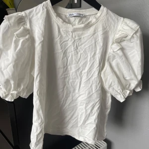 Vit puffärmad topp från Zara - Säljer en vit topp från Zara med puffiga korta ärmar och volangdetaljer vid axlarna. Toppen har rund halsringning och är gjord i bomull, perfekt för en trendig och stilren look.