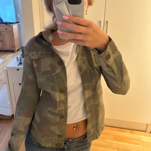 Camo militärinspirerad jacka med stjärndetaljer - Säljer en grön camouflagejacka med knappar och coola stjärndetaljer på ena bröstfickan. Jackan har klassisk krage, två bröstfickor och är långärmad. Står ingen storlek men jag skulle säga xs.