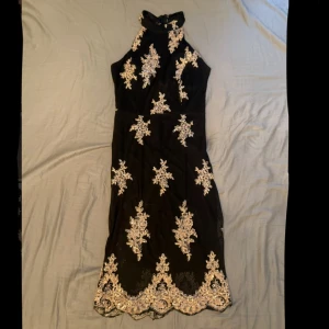 Svart och beige klänning - Säljer en svart klänning med vacker beige-aktig brodyr och hög krage. Klänningen är ärmlös och har figurnära passform med fina detaljer längs nederkanten och över hela plagget. Har endast använts ett par gånger och har några fel som syns på bilderna ovan. Skriv vid frågor om måtten.