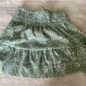 Grön blommig kjol från Gina Tricot - Supersöt grön kortkjol från Gina Tricot med vitt blommönster och smock i midjan. Kjolen har volangdetaljer och är perfekt för vår och sommar. Lätt och luftig modell som är enkel att styla med en enkel topp. 💗