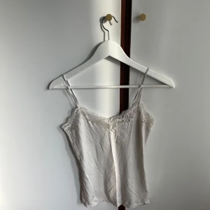 Vit spetsdetaljerad linnetopp från Lindex. - Säljer en söt vit linnetopp från Lindex med tunna axelband och fina spetsdetaljer längs urringningen.