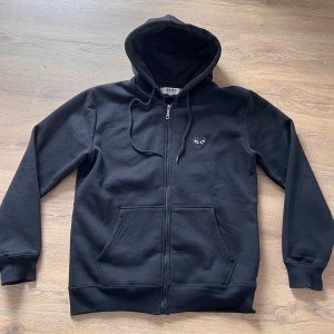 Svart hoodie med dragkedja från Comme des Garçons Play - Svart hoodie med dragkedja och huva från Comme des Garçons Play. Tröjan har ett litet hjärta med ögon broderat på bröstet och en stor hjärtdetalj på ryggen. Praktiska fickor framtill och justerbar dragsko i huvan.