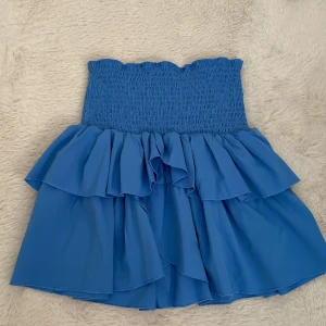 Blå volangkjol från Neo Noir - Supersöt blå kjol från Neo Noir med smockad midja och dubbla volanger. Kjolen är kort och har en luftig känsla, perfekt för varma dagar. Enkel att matcha med både toppar och t-shirts.