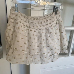 Vit kjol med guldprickar och volang - Supersöt vit kjol med guldiga prickar och bred smock i midjan. Kjolen har volangdetaljer och är gjord i ett lätt, transparent material som ger en luftig känsla. Perfekt för dig som vill sticka ut lite extra!