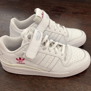 Vita Adidas sneakers med rosa logga - Säljer ett par fräscha vita Adidas sneakers med klassisk design och rosa detaljer. Skorna har snörning och kardborreband. Har använts ett fåtal gånger men skicket är så gott som nytt. Se två sista bilder för spår av användning