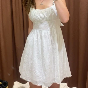 Vit kortklänning med knyt i ryggen från Gina tricot - Superfin vit klänning med smala axelband och söt knytning i ryggen. Klänningen har broderade detaljer. Jättefin till sommaren och skolavslutning. Använd en gång. Strl 42 men passar bra på mig som oftast har 36!