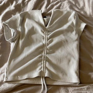 Beige ribbad topp med knyt - Säljer en beige ribbad topp med korta ärmar och v-ringning. Toppen har en snygg knyt- och rynkdetalj framtill som gör att du kan justera längden. Perfekt till jeans eller kjol för en avslappnad look. ca 60 cm i midjan