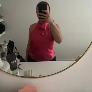 Rosa halterneck topp i satin - Säljer en snygg rosa halterneck topp i satin med öppen rygg. Toppen har en draperad halsringning och är ärmlös, perfekt för att sticka ut. Sitter tight upptill och har en elegant glansig finish.