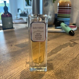 Laura Mercier Almond Coconut Milk Eau de Toilette - En elegant parfym från Laura Mercier med doft av mandel och kokosmjölk. Flaskan är rektangulär i genomskinligt glas med silverfärgad spraykork. Parfymen har en ljusgul vätska och en stilren etikett framtill.