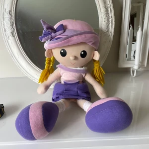 Mini Rag Dolls - Söt mjukdocka med lila hatt och klänning. Dockan har blont hår i flätor och stora, mjuka fötter. Perfekt som en kramgo vän eller dekoration i barnrummet. Mäter cirka 55cm