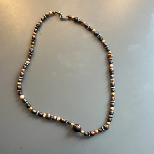 Brunt pärlhalsband med silverdetaljer - Säljer ett handgjort halsband med bruna och beige pärlor i olika former, varvat med små silverfärgade detaljer. Halsbandet har ett klassiskt spänne och en större rund pärla i mitten. 