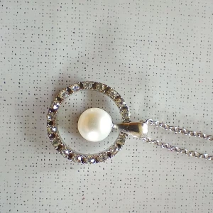 Silverfärgat halsband med pärla - Halsband i silverfärg med ett runt hänge med stenar och en vit pärla i mitten. Kedjan är tunn och justerbar. Mycket fint skick