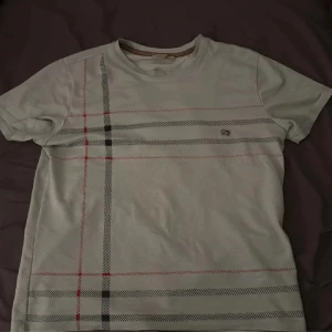 Ljusblå Tshirt från burberry - Säljer en vit t-shirt från burberry diskret rutigt mönster i svart, grått och rött på framsidan. T-shirten har rund halsringning och korta ärmar. det står M på den men den passar bara I S och XS